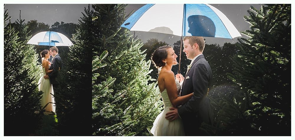 A couple embraces under an umbrella in a rainy forest. The woman wears a white dress.