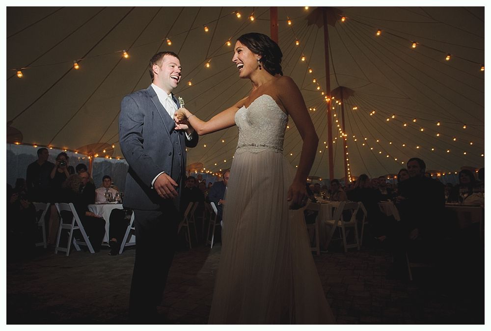 Bride and groom laughing while dancing at a wedding reception under string lights.