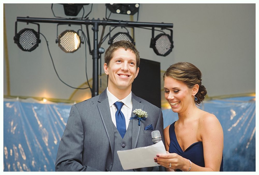 Smiling man and woman at a wedding, he in a suit, she in a dark dress, holding paper, with stage lights.