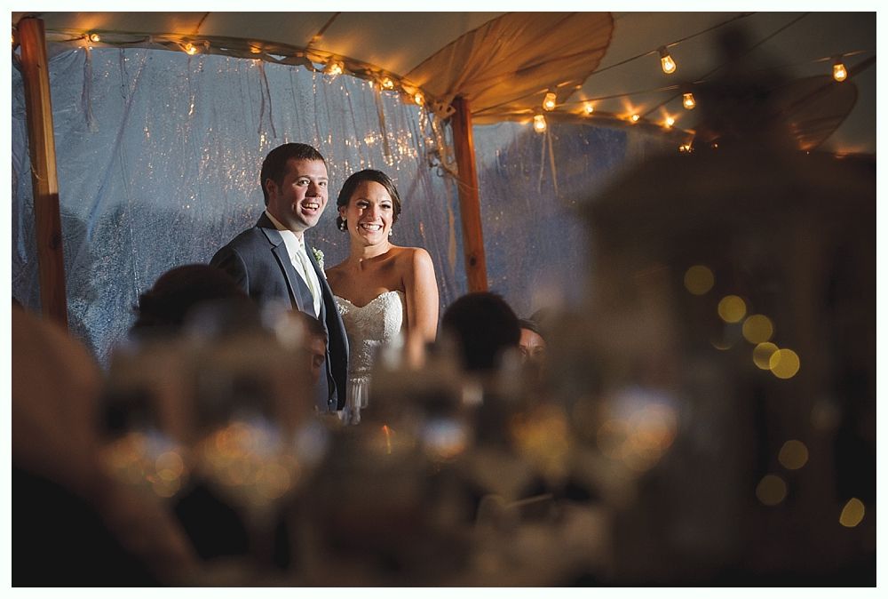 Couple at a wedding, smiling under string lights. The woman wears a white strapless dress, the man a suit.
