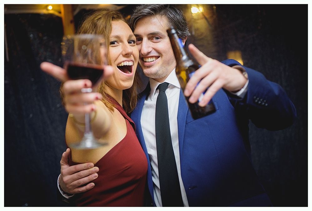 Woman in red dress and man in blue suit celebrate, holding wine and beer. Both smiling.