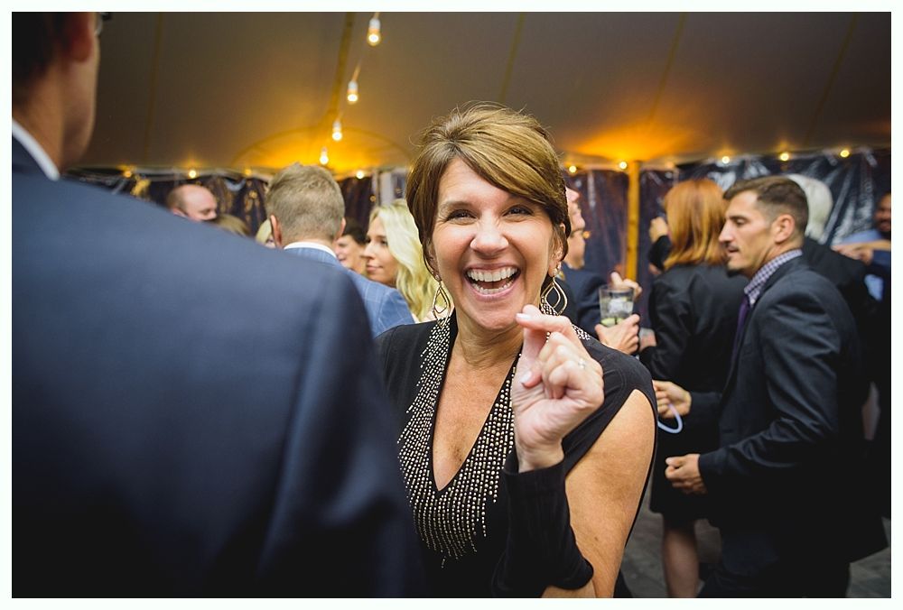 Woman smiling broadly at a party, raising her hand with a small gesture. Partygoers in background.