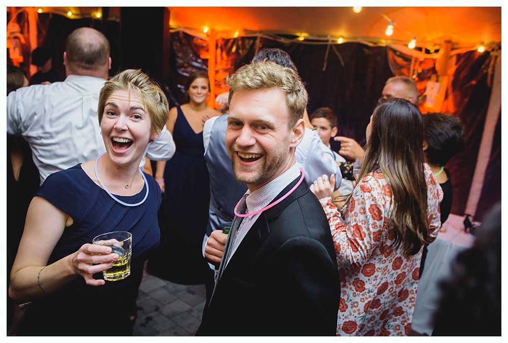 Two people smiling and laughing at a party, holding drinks, with string lights and people dancing in the background.