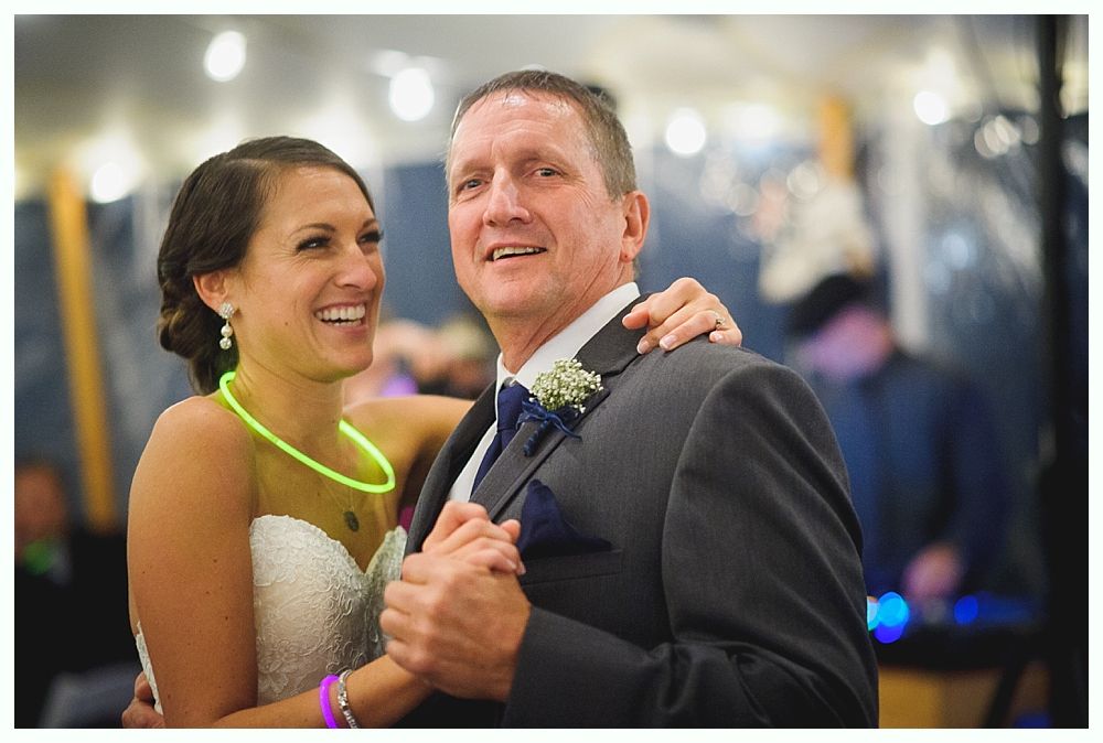 Bride and man dance at reception, smiling. Bride in white dress, neon necklace. Man in gray suit.