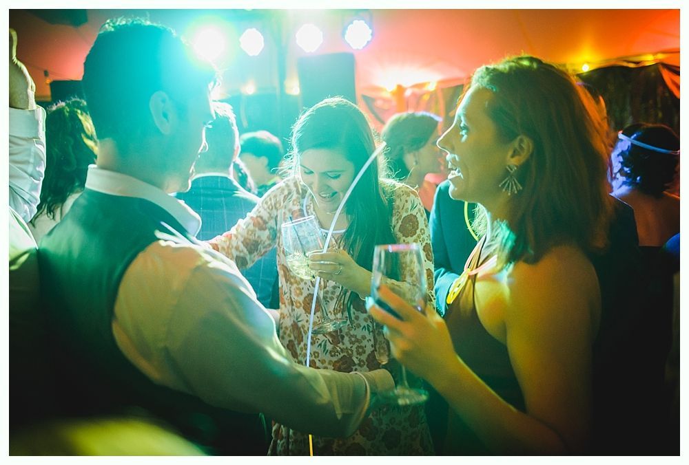 People laughing and talking at a party, holding drinks. Bright lights illuminate the lively scene.