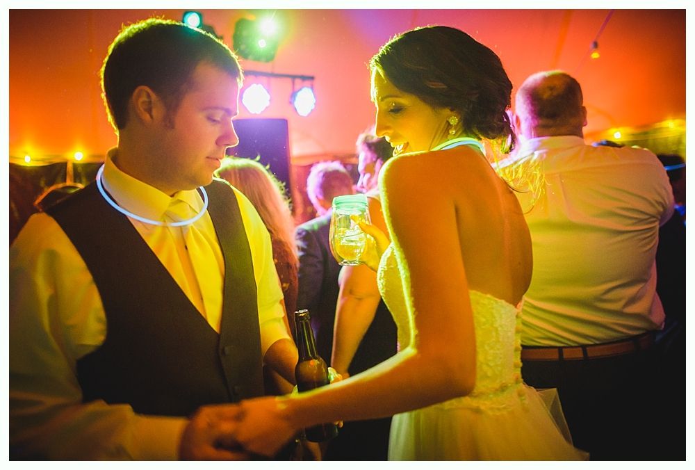 Couple dancing at a wedding reception, wearing glow necklaces, surrounded by guests and colorful lights.