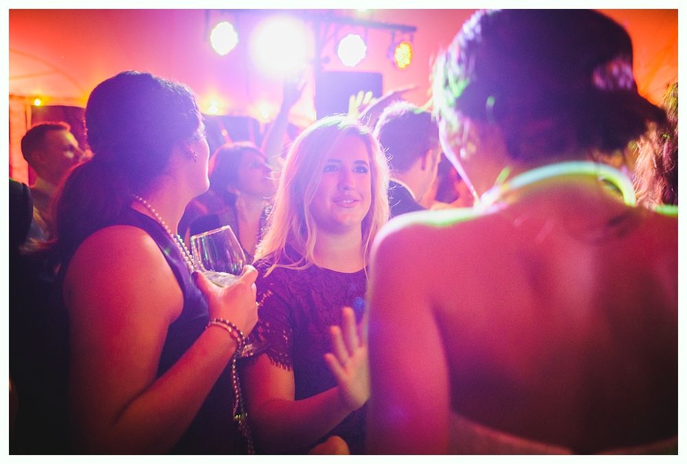 People dancing at a party with a woman in the center smiling. Colorful lights illuminate the room.