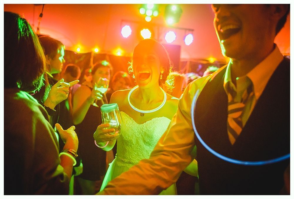 People celebrating at a party with colorful lights. Woman in a dress laughs, holding a drink. Man in vest laughs, too.