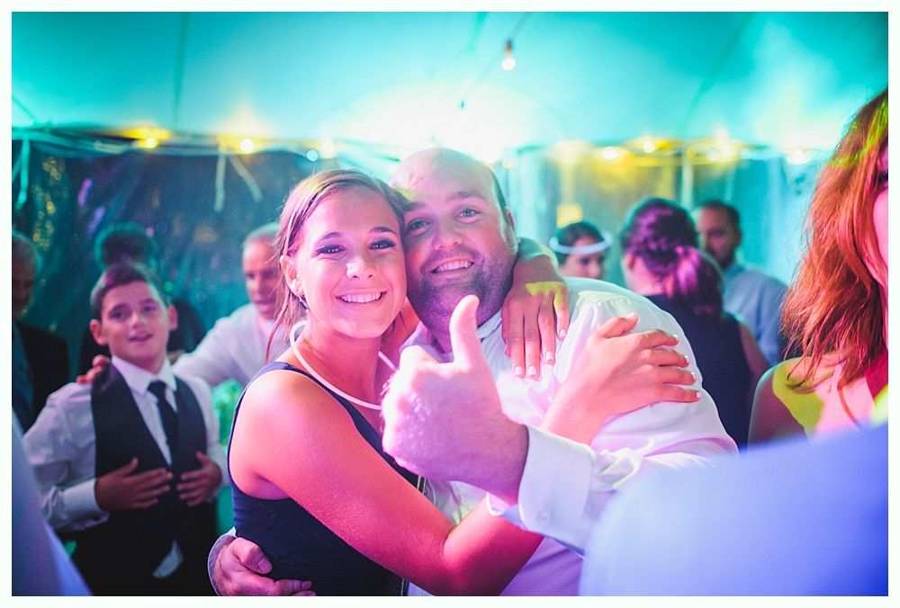 Couple hugging, smiling, at a party, man gives a thumbs up. Lit with blue and pink light.