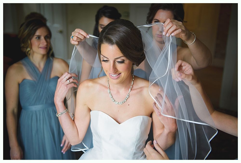 Bride in white strapless dress has veil adjusted by bridesmaids in blue dresses. Warm lighting.
