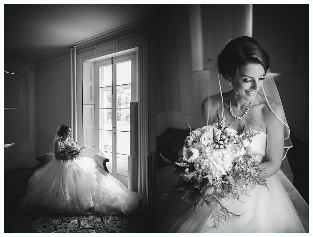 Bride in a white gown holds bouquet, smiling near a window, black and white photograph.