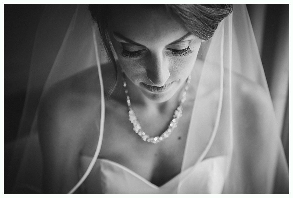 Bride looking down, wearing a veil and beaded necklace. Black and white photo.