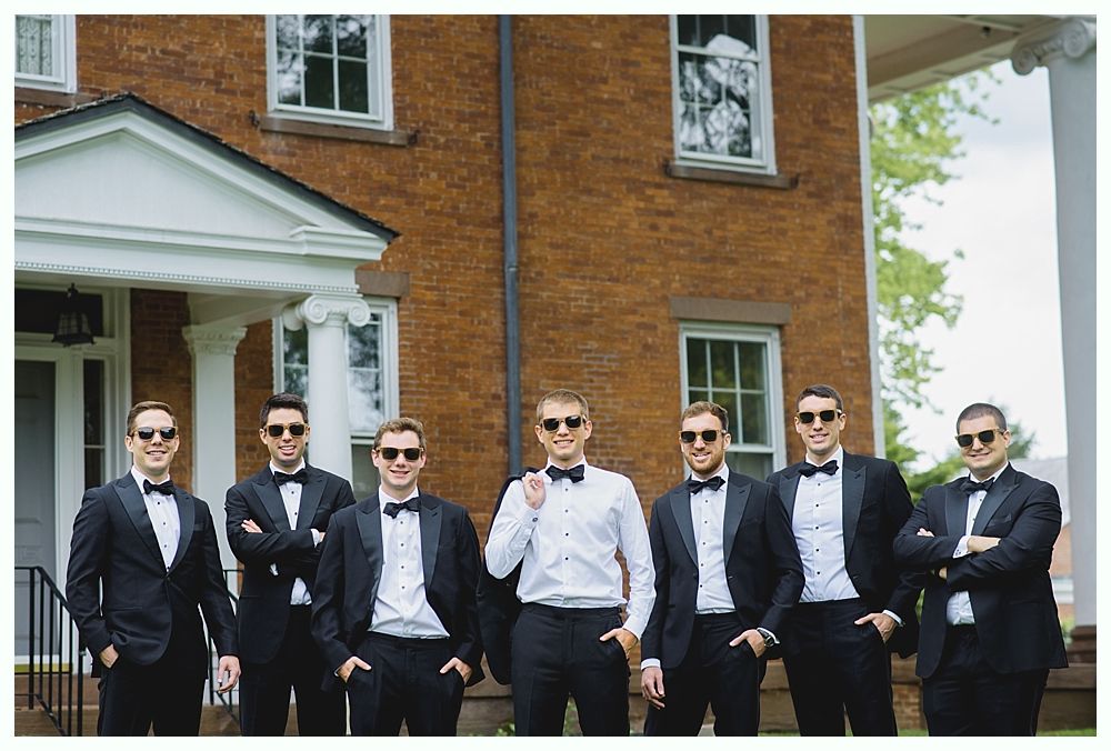 Men in tuxedos pose outside a brick building, wearing sunglasses and smiling.