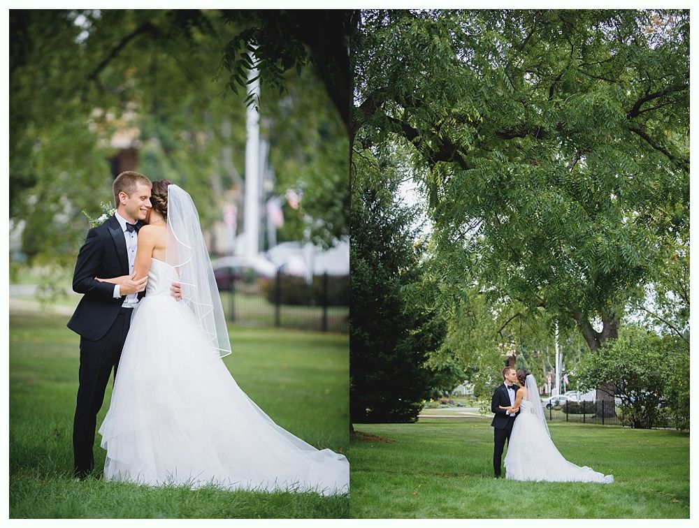 Bride and groom pose on a grassy lawn under a large tree, she in a white gown and veil, he in a black tuxedo.