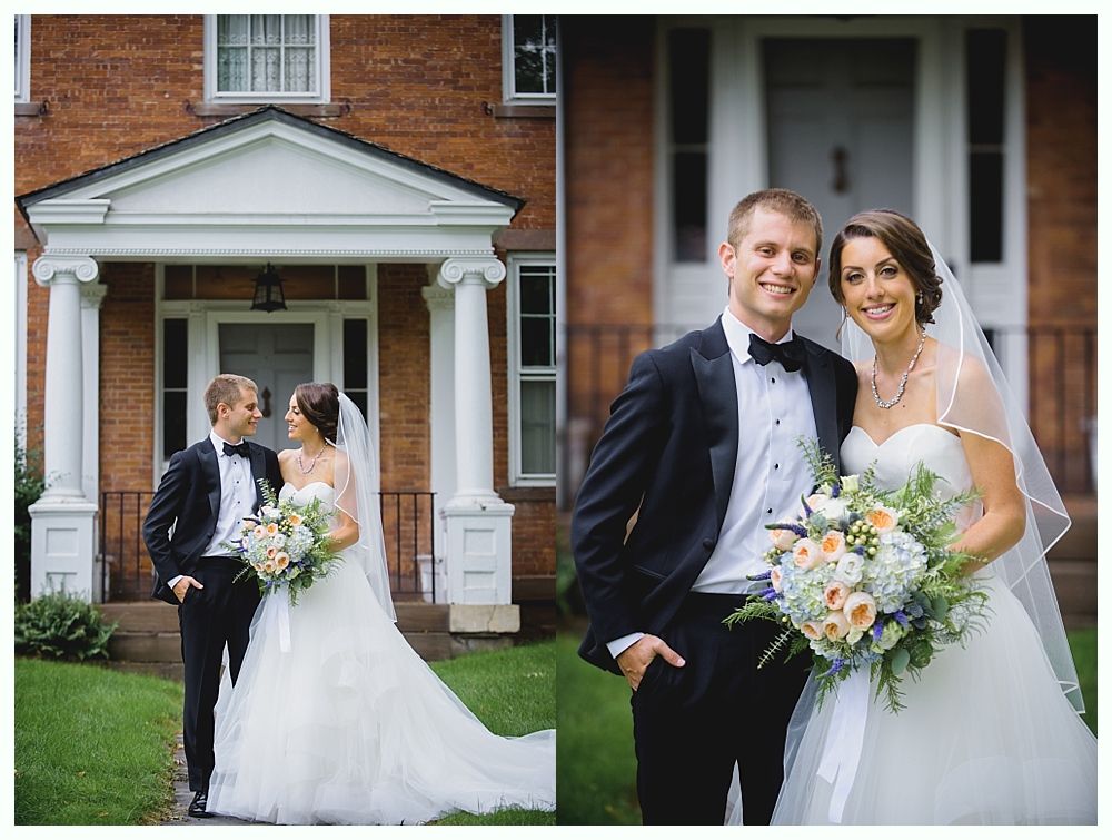 Bride and groom pose in front of a brick building with white columns and doorway. They are smiling and holding flowers.