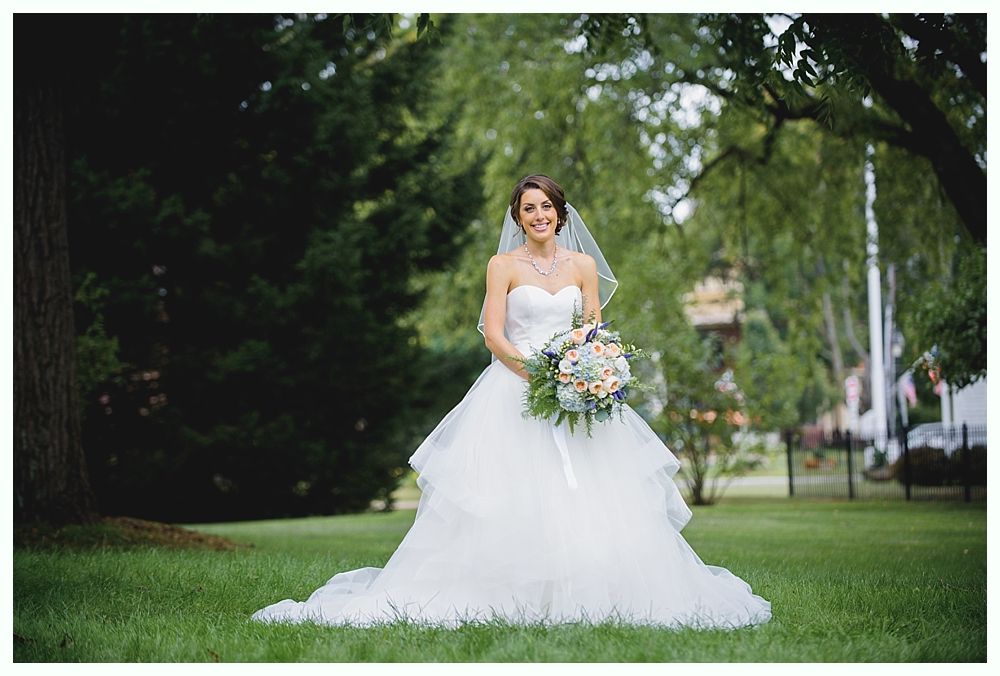 Bride in a white strapless gown holding a bouquet, standing in a grassy area with trees.
