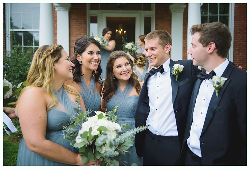 Wedding party poses in front of a brick building. Bridesmaids in blue dresses, groomsmen in tuxedos, smiling.