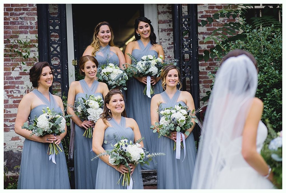 Bridesmaids in blue dresses with bouquets smile at the bride, standing in front of a brick building.