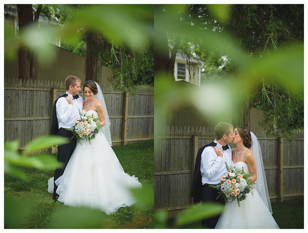 Bride and groom kissing, standing in a yard. She wears a white dress and veil, he wears a tuxedo. Fence and tree in the background.