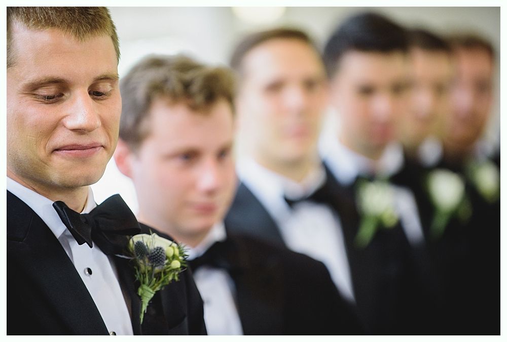 Groom and groomsmen in black tuxedos, waiting at the altar. Focus on groom looking down, emotional.