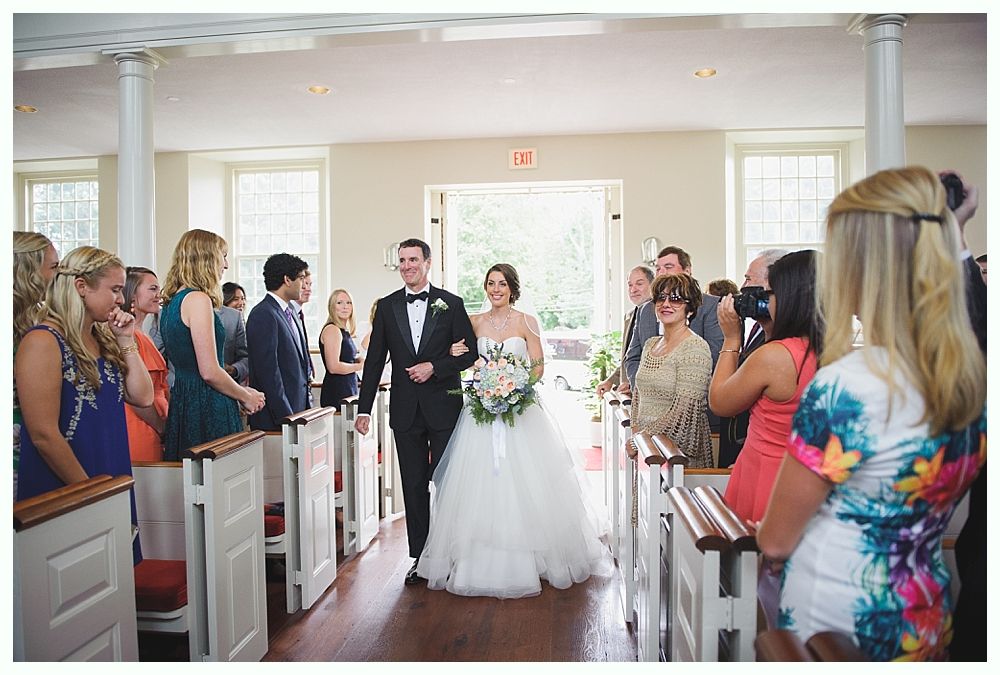 Bride and groom walk down aisle after wedding ceremony, smiling. Guests watch from pews in a white-walled church.
