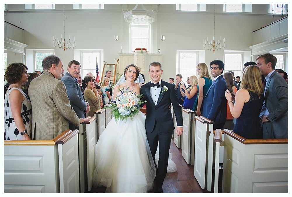 Newlyweds exiting a white church, smiling, holding hands. Guests in pews. Bride in white gown, groom in tuxedo.