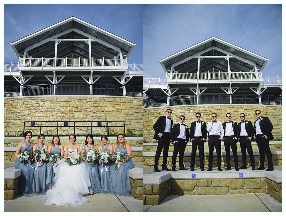 Wedding party posing in front of a building with a stone facade. Bridesmaids in blue dresses, groomsmen in black tuxedos.