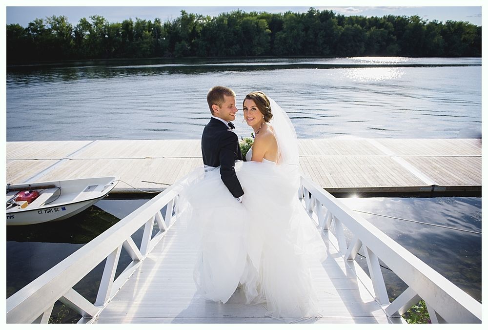 Bride and groom on a white dock overlooking water. Bride in white gown, groom in tuxedo, looking back at the camera.