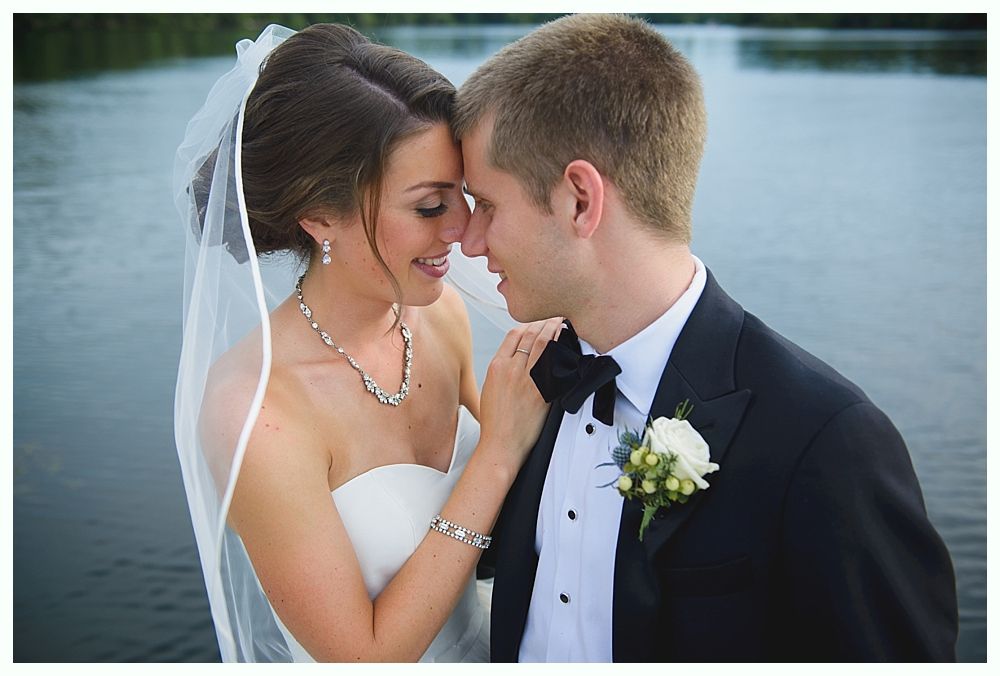 Bride and groom in formal wear touch foreheads. She smiles, wearing a veil and jewelry. River in background.
