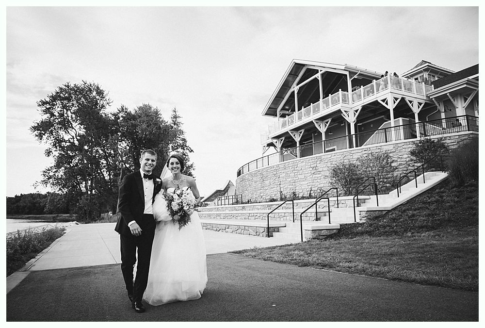 Bride and groom walking on a paved path in front of a large venue. Black and white photo.