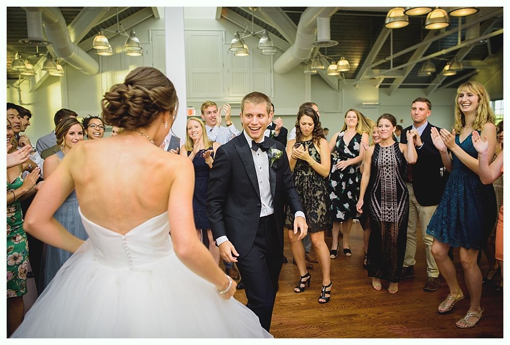 Bride and groom dance at a wedding reception with guests clapping; bright, indoor setting.