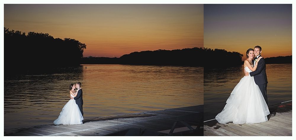 Couple embracing on a lake shore at sunset. She wears a white wedding dress, he wears a suit. Orange and purple sky.