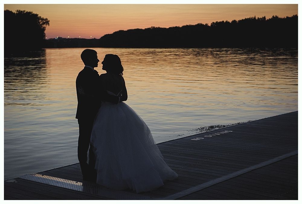Couple in silhouette on dock at sunset, embracing, water and trees in background.