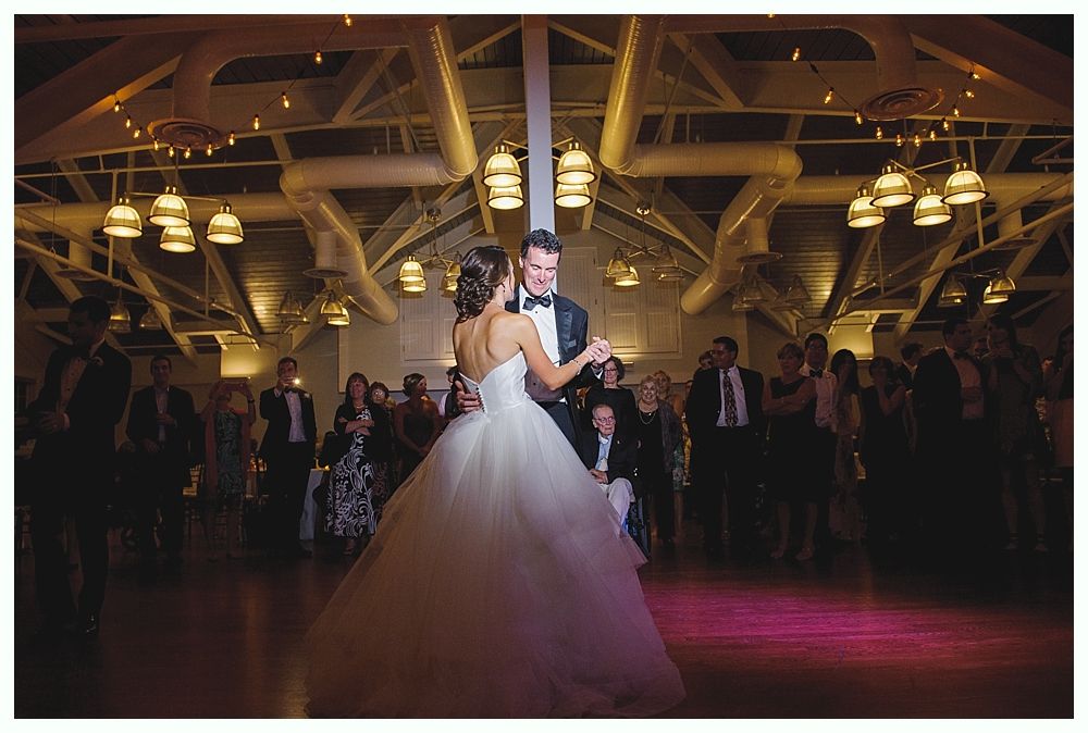 Bride and groom dance at a wedding reception. They are center, surrounded by guests in a large hall with ambient lighting.