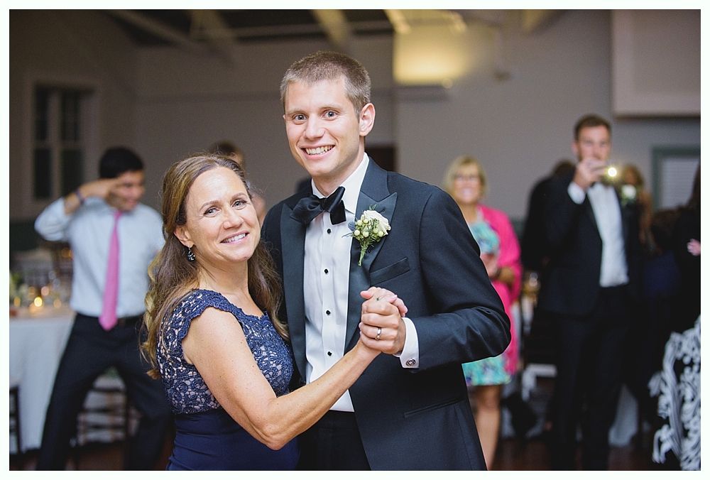 Man in tuxedo dances with woman in blue dress at a wedding reception. People in background.