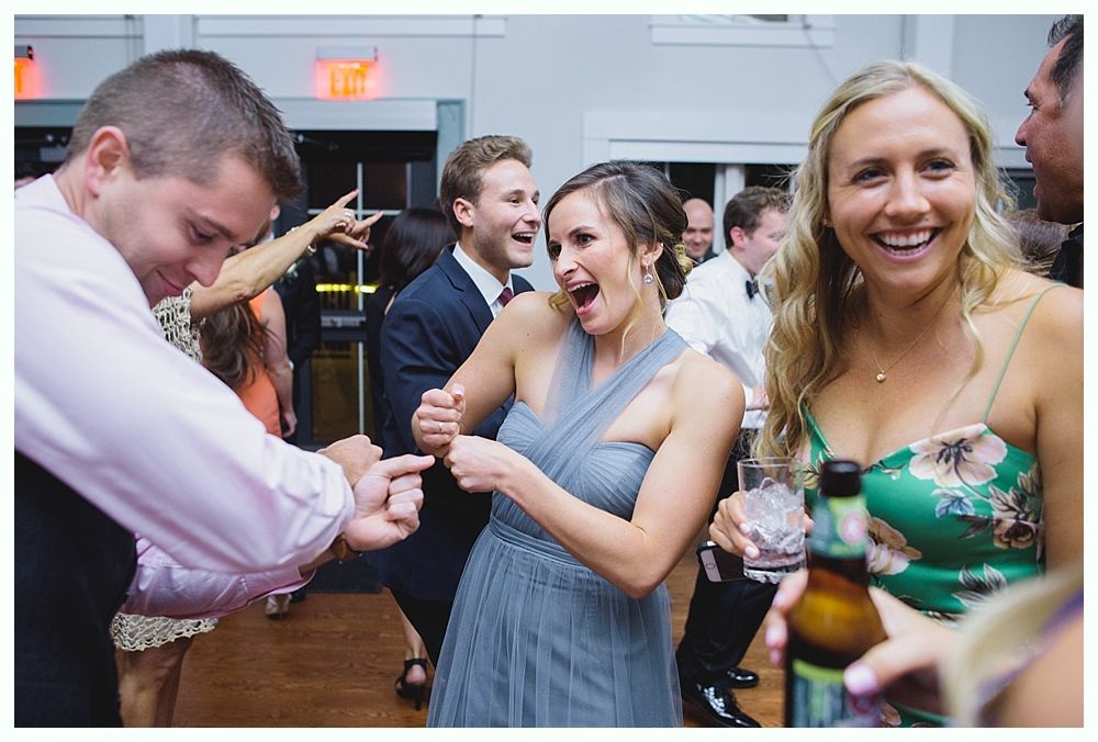 People dancing at a wedding reception; woman in gray dress points at man, all smiling and laughing.
