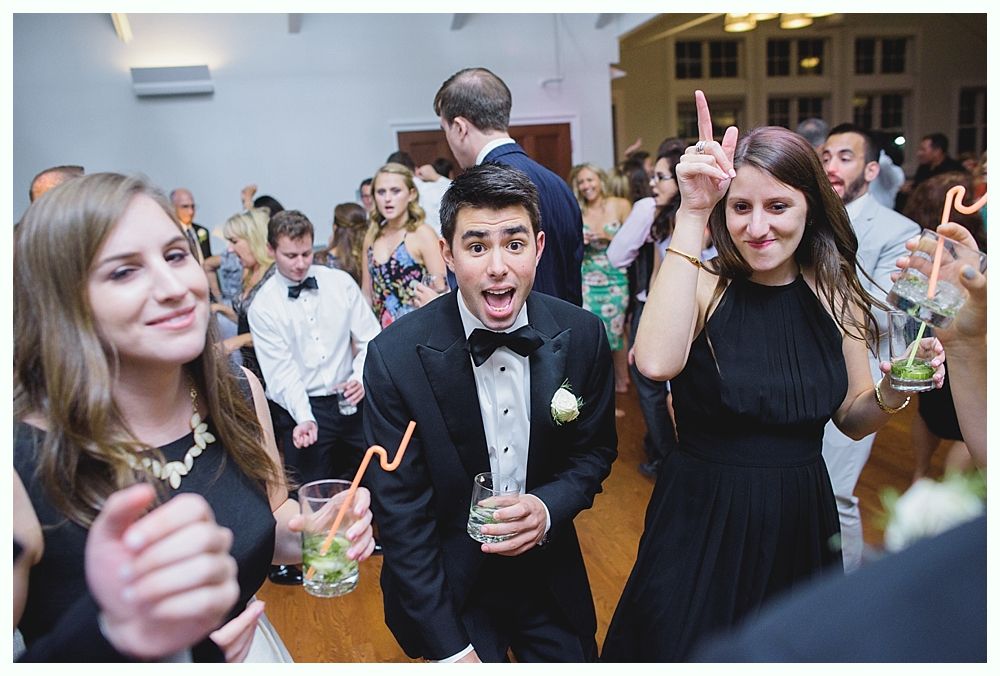 People dancing at a party; man in tuxedo makes a silly face, woman raises fingers, drinks in hand.