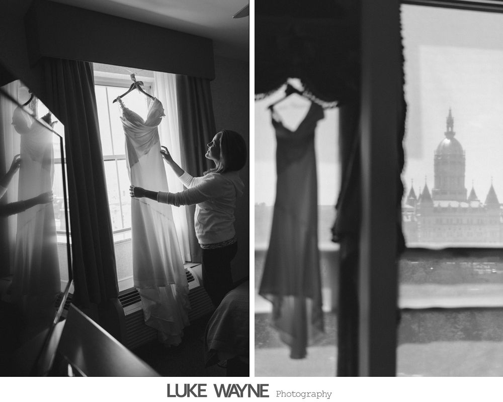 Wedding dress hanging in a window; woman adjusts it; cityscape visible outside. Black and white.
