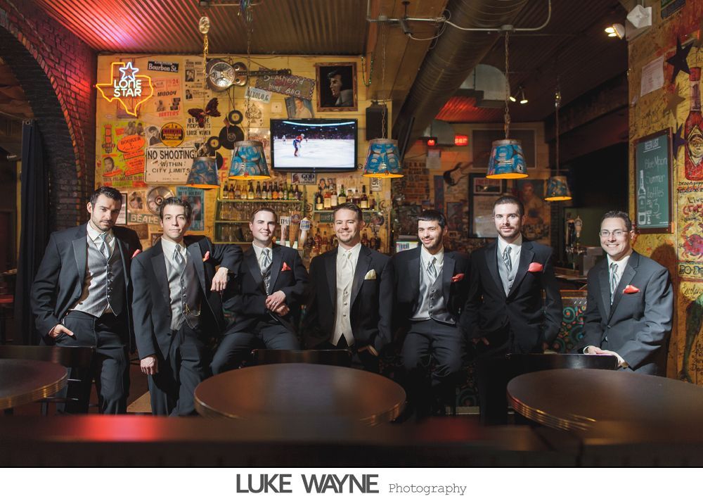 Groomsmen in suits pose at a bar. Dark setting with a festive vibe, men smiling at the camera.