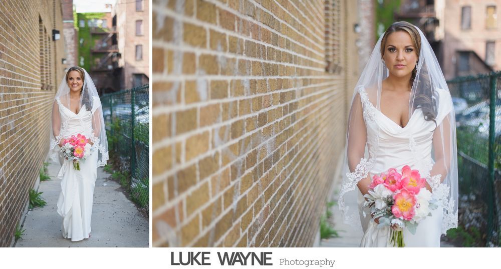 Bride in a white gown and veil holding flowers, walking in an alleyway.