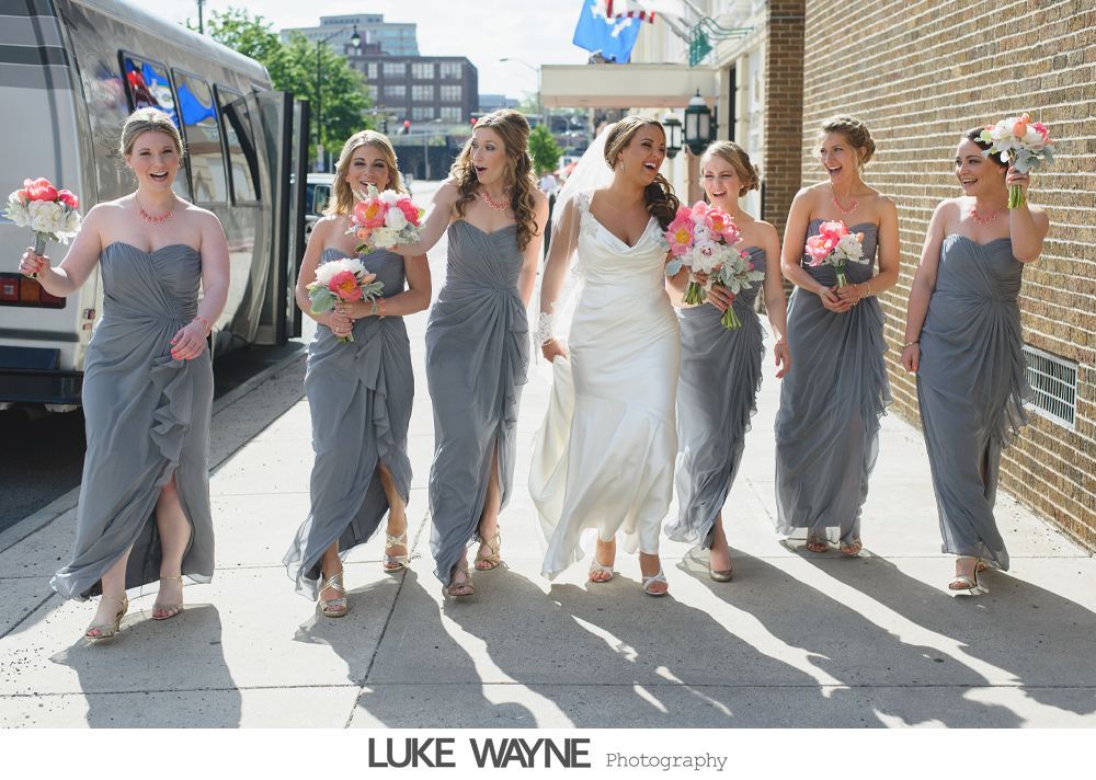 Bride and bridesmaids in gray dresses walking down a sidewalk, smiling, holding bouquets.