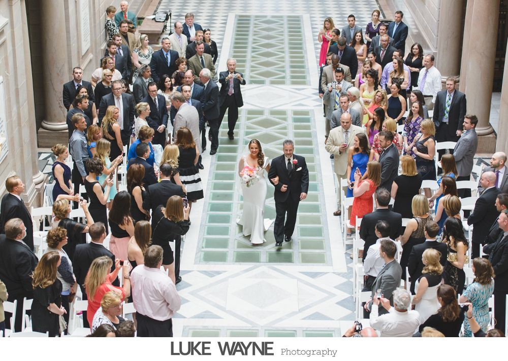 Bride escorted down aisle by father, guests watching, indoor ceremony.