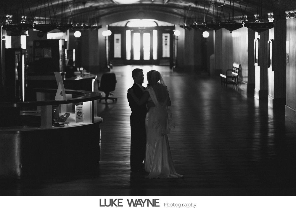 Bride and groom embrace in a dimly lit hallway; the dark tones contrast with the bright doorway at the end.