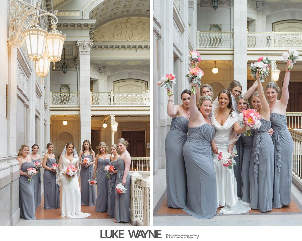Bride and bridesmaids in gray dresses pose inside a building, holding bouquets and celebrating.