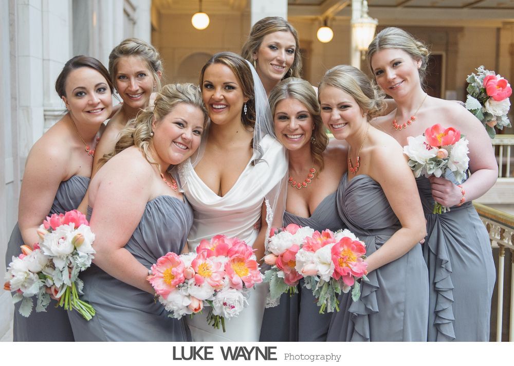Bride and bridesmaids in gray dresses pose, holding coral and white bouquets.