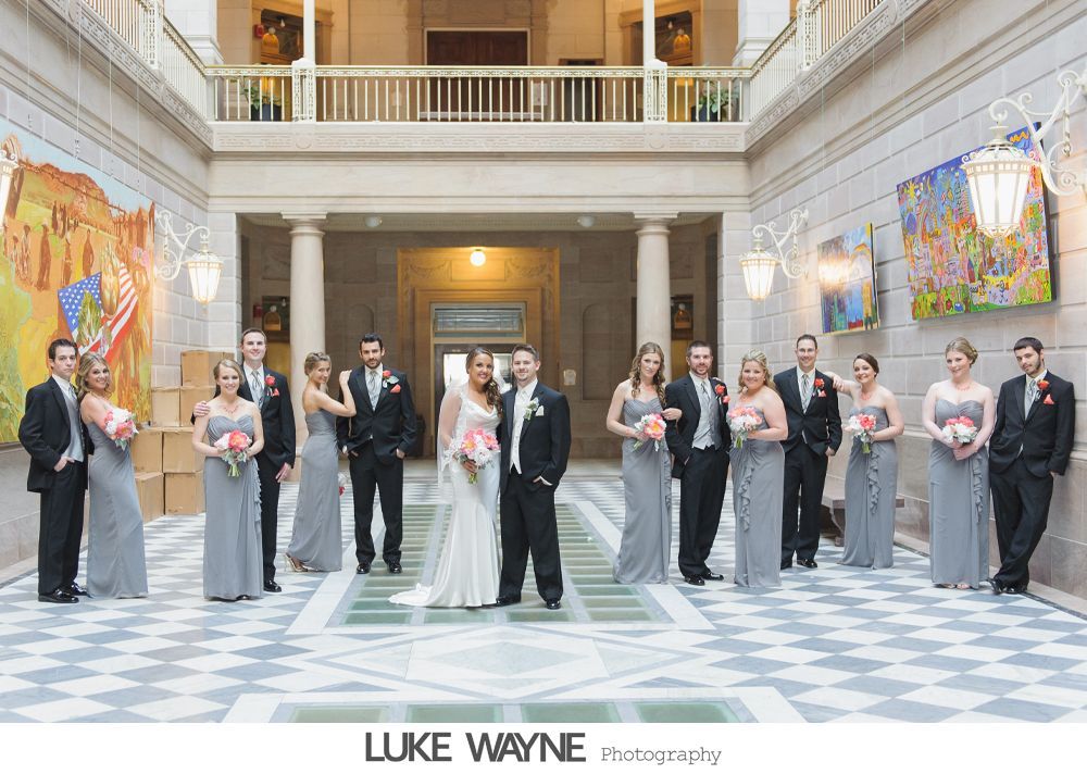 Wedding party poses in a grand hall with bride and groom centered; bridesmaids in gray gowns, groomsmen in black suits.