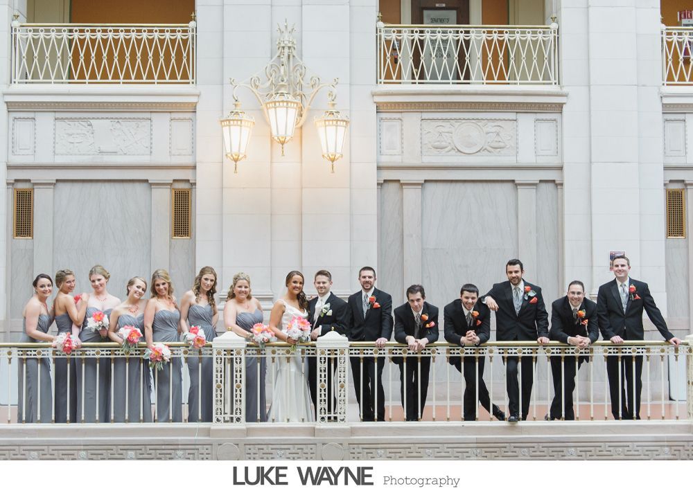 Wedding party on balcony, women in gray dresses with bouquets, men in black suits, ornate building.