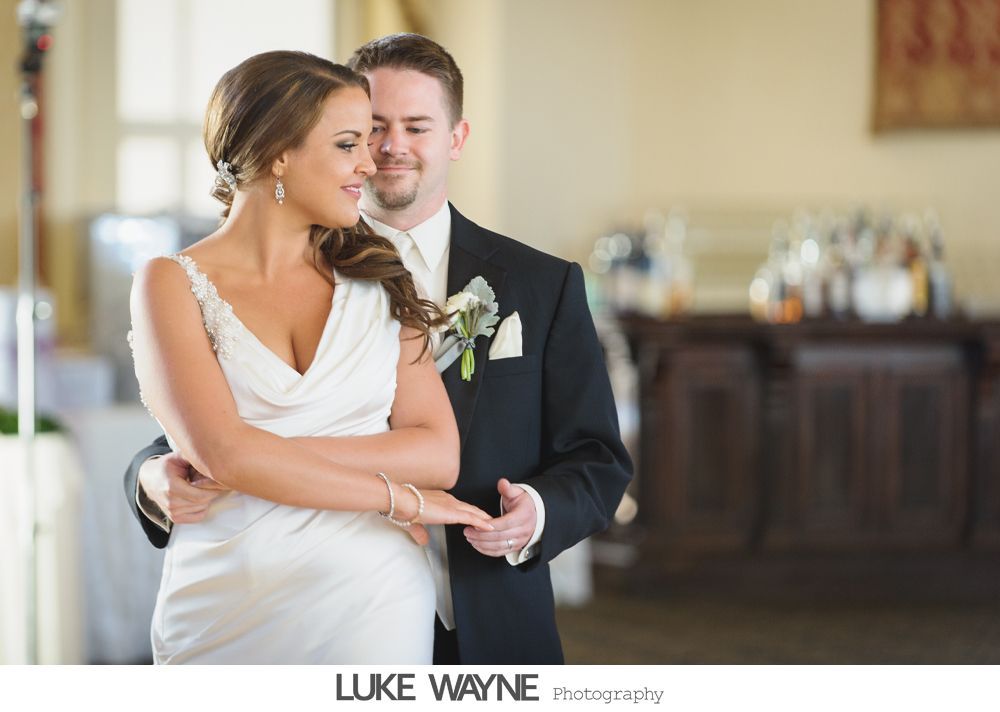 Bride and groom embracing indoors. The bride wears a white dress and the groom a black suit, smiling.