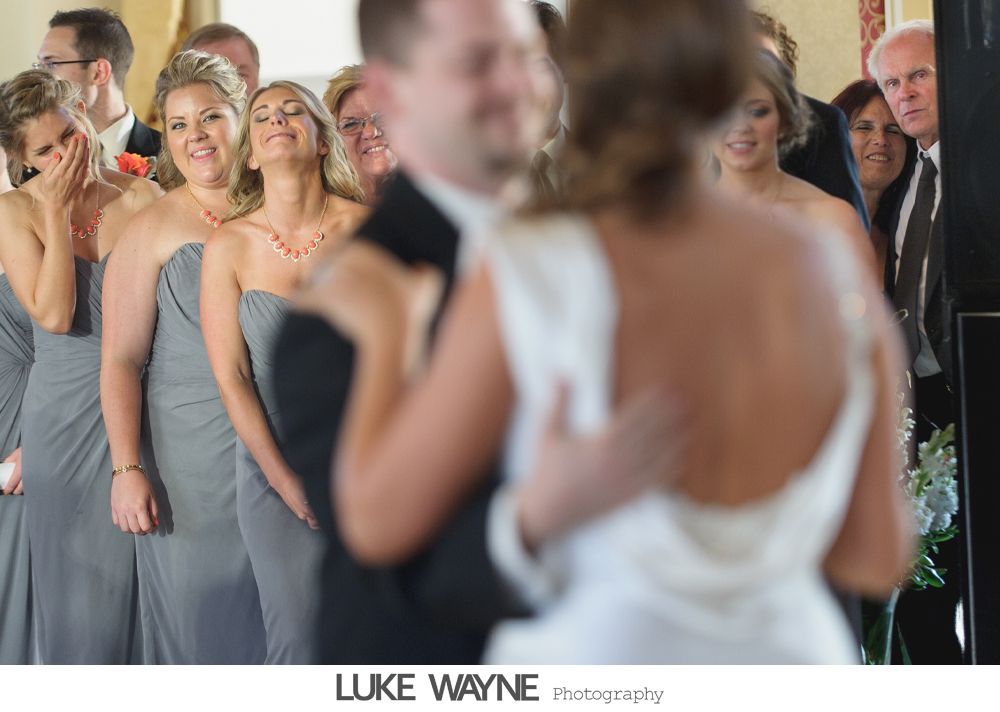 Wedding party watches the bride and groom dance; bridesmaids in gray dresses, all smiling.