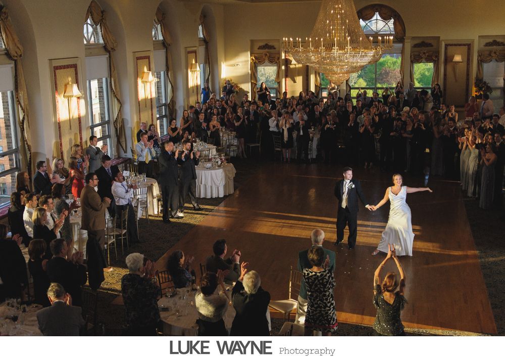 Bride and groom dance at reception; guests applaud. Ballroom with chandelier, sunlight through windows.